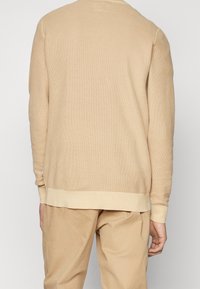 Beige pletený svetr s texturovaným vzorem, žebrovanými manžetami a lemem. Hladký zadní design, minimální detaily a boční rozparky ve spodní části.