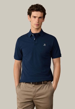 Junger Mann trägt ein marineblaues Polohemd mit Logo und beige Hose, steht mit den Händen in den Taschen vor einem einfarbigen Hintergrund.