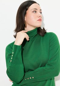 Pull à col roulé vert en texture côtelée avec des détails de boutons dorés aux manches. Tissu lisse avec une silhouette ajustée.