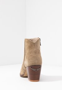 Karmelowe zamszowe botki za kostkę o wyrazistym kształcie, z teksturowanym detalem z tyłu oraz drewnianą podeszwą z akcentem w postaci srebrnych skrzydeł.
