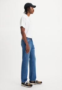 T-shirt blanche à manches courtes et jeans en denim bleu, associés à une casquette noire et des baskets multicolores. Le modèle se tient de côté devant un fond blanc.