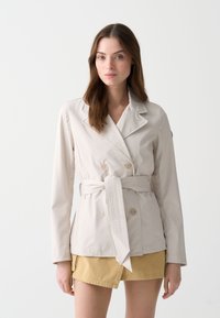 Veste beige à double boutonnage avec un large col, ceinture à nouer et détails de boutons. Texture lisse, manches longues et coupe ajustée.