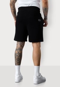Shorts noirs en coton avec une taille élastique et des poches latérales, présentant des détails de logo discrets. Portés avec des baskets blanches et des chaussettes.