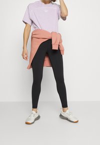 Jasnofioletowy t-shirt z białymi akcentami, czarne legginsy i różowy sweter przewiązany w talii. Białe sneakersy z szarymi podeszwami i pomarańczowym wykończeniem.
