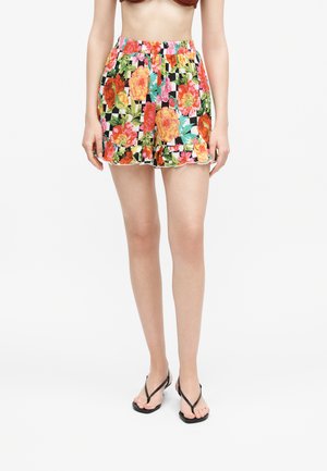 Žena má na sebe živé šortky s kvetinovým vzorom a nariaseným lemom a čierne žabky, stojí pred jednoduchým bielym pozadím.