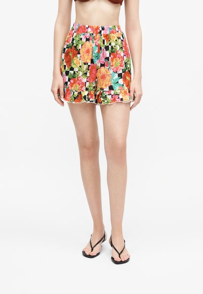Femme portant un short à imprimé floral vif avec un ourlet volanté et des sandales noires en forme de tongs, debout devant un fond blanc uni.