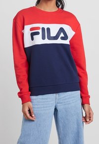 Personne portant un sweat-shirt FILA rouge, blanc et bleu marine, associé à un jean bleu clair, debout devant un fond clair uni.