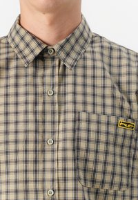 Close-up of a person's torso wearing a tan plaid shirt with brown and blue lines, featuring a chest pocket and a yellow logo.