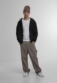 Sort zip-up hoodie over en hvid T-shirt, beige cargo bukser og en brun beanie-hat med et logo. Hvide sneakers fuldender outfittet.