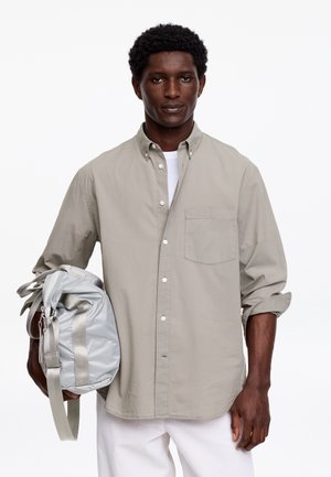 Vyras, vilkintis smėlio spalvos marškinius su sagutėmis ir baltas kelnes, laikantis sidabro pilkos spalvos sportinę rankinę kairėje pusėje baltame fone.