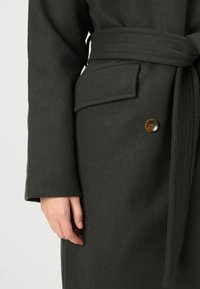 Dypgrønn ullfrakk med belte i midjen, sidelommer og store knapper. Stoffet har en teksturert overflate og en skreddersydd passform.