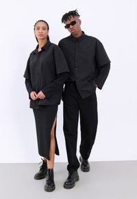 Una camisa negra de algodón oversize en una mujer, combinada con una falda negra con una abertura lateral. Un hombre lleva pantalones negros y botas de combate.