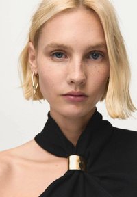 Top de tela negra con diseño anudado y detalle de herrajes dorados. Modelo con cabello rubio corto y piel clara con pecas.