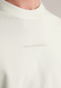 Biały bawełniany t-shirt z prążkowanym kołnierzykiem i subtelnym szarym haftem z napisem "marcofoldenim" na klatce piersiowej. Prosty, minimalistyczny design.