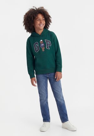 Παιδί με κατσαρά μαλλιά που χαμογελά, φοράει πράσινο φούτερ με κουκούλα με λογότυπο αρκούδας και μπλε τζιν, στέκεται μπροστά σε απλό φόντο.