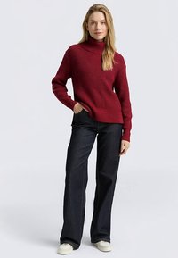 Red ribbed knit turtleneck sweater with long sleeves, worn with dark denim flared pants and white sneakers. Sweater has a relaxed fit.