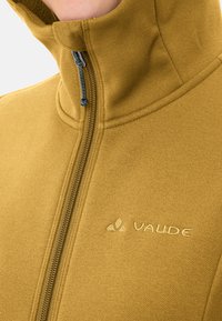 Gevoerd fleece jack in mosterdgeel met een gladde textuur. Beschikt over gestikte details en een klein geborduurd logo op de borst.