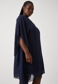 Robe fluide bleu marine en tissu léger, à coupe ample avec des manches drapées et un ourlet asymétrique.