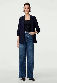 Mornarska blazerica preko črne brezrokavne majice, v kombinaciji z širokimi modrimi kavbojkami. Obleka se ponaša z sproščenim krojem in gladko teksturo.