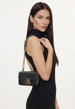 Femme aux longs cheveux foncés portant une robe noire sans manches, tenant un sac à main en cuir noir avec une bandoulière chaîne dorée sur l'épaule.