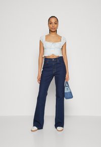 Top corto di un blu chiaro con pizzo, abbinato a jeans a zampa d'elefante scuri a vita alta. Tiene in mano una piccola borsa di denim. Sneakers bianche.