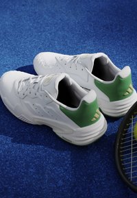 Scarpe sportive bianche con dettagli verdi sul tallone su una superficie blu accanto a una racchetta e una palla da tennis.