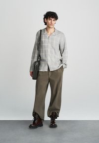 Světle šedá kostkovaná košile na knoflíky, béžové volné kalhoty, tmavě hnědé šněrovací boty a černá kabelka přes rameno, na jednobarevném pozadí.