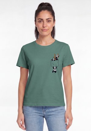 Vrouw draagt groen T-shirt met klein zakontwerp van drie speelse panda's die klimmen en zitten met bamboestokjes.