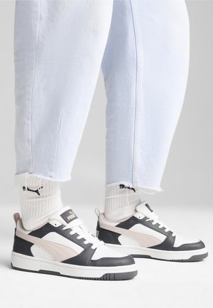 Fødder iført lyseblå jeans, hvide Puma-strømper og sorte, hvide og beige Puma-sneakers med snørebånd på en hvid gulv.