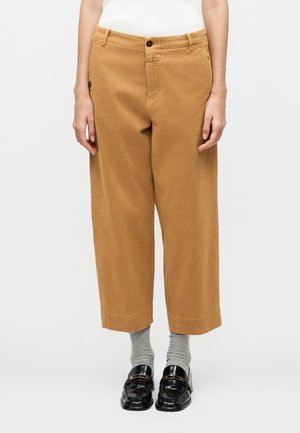 Άτομο που φοράει καφέ παντελόνι με φαρδιά κοντή γραμμή, γκρι κάλτσες, μαύρα μοκασίνια και λευκό αμάνικο τοπ, στέκεται μπροστά σε απλό φόντο.