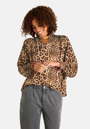 Woman smiling down, wearing a long-sleeve leopard print blouse with pink-trimmed cuffs and gray high-waisted pants.