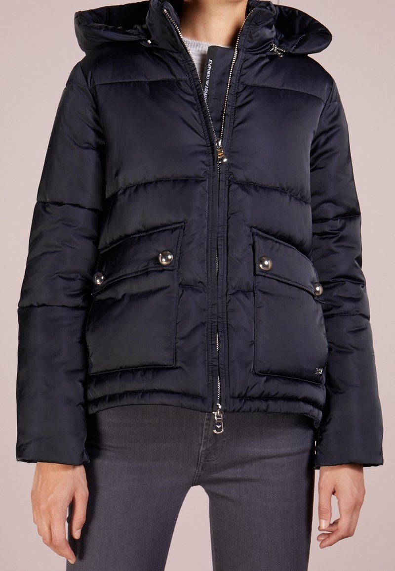 Marinblå puffjacka med huva, som har blixtlås, två framfickor med tryckknappar och en quiltad textur. Silverfärgade detaljer.