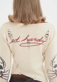 Beige gebreide trui met verwoeste delen, geborduurde "ed hardy" logo en doodskopontwerpen op de mouwen. Losvallende pasvorm met een ruwe zoom.