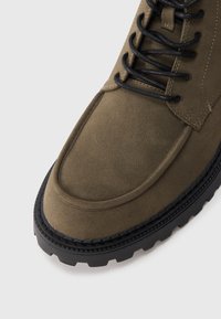 Bota de ante verde oliva con cordones negros, punta redondeada y suela gruesa de goma negra con un dibujo de tracción.