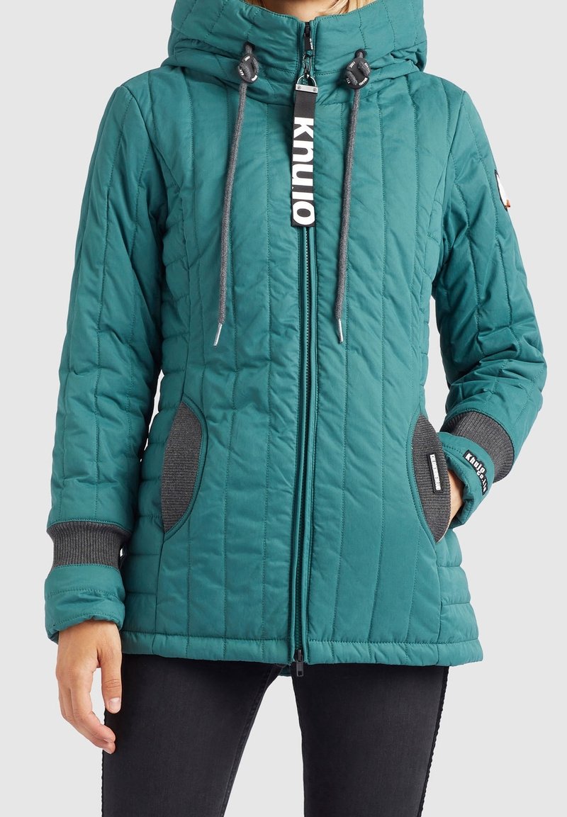 Veste matelassée turquoise avec capuche, fermeture éclair à l'avant, poignets en tricot gris et accents de poche, avec tirette de fermeture éclair marquée "knullo", portée avec un pantalon noir.