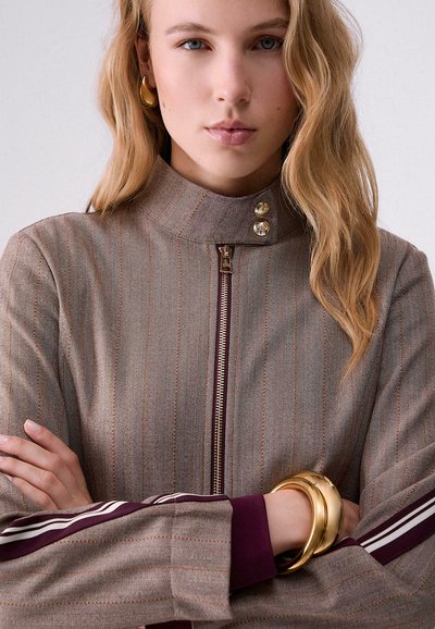 Jeune femme aux cheveux blonds portant une veste rayée à col montant avec fermeture éclair, des boucles d'oreilles en or et deux bracelets en or croisant ses bras.