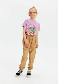 Lilla T-shirt med blomstermotiv og tekst; lysebrune bukser med elastisk talje; sort-hvide sneakers med snørebånd; pige der smiler, med kort hår.