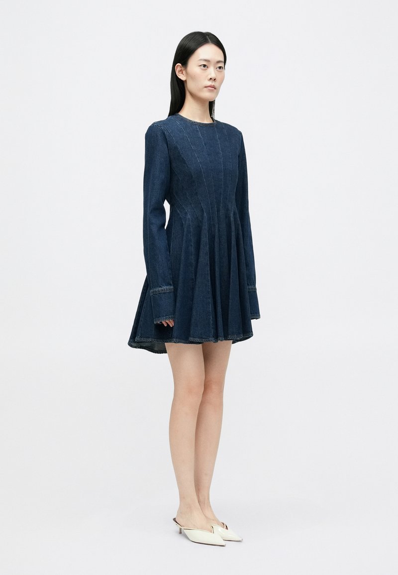 Robe en denim bleu foncé avec des manches longues, un buste ajusté et une jupe évasée. Présente des coutures visibles et un col rond.