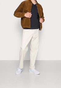 Osoba ubrana w brązową kurtkę sztruksową na czarnej koszuli, kremowe spodnie i białe trampki, stojąca na jednolitym jasnym tle.