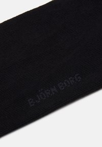 Crna čarapa izrađena od teksturiranog pletiva, s diskretnim tiskanim logotipom u tamnijoj nijansi sa strane.