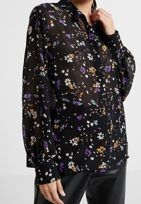 Blusa nera trasparente con motivo floreale in viola, bianco e arancione. Presenta una chiusura a bottoni sul davanti e maniche lunghe a sbuffo. Tessuto morbido.