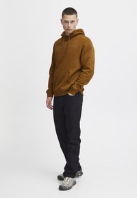Un sweat à capuche marron avec une poche kangourou et le mot "AMSTERDAM" en relief sur le devant, associé à un jean noir et des chaussures de sport grises.