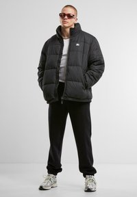 Sort puffervest med høj krave og lynlås, båret over en hvid skjorte. Kombineret med sorte sweatpants og sneakers med multicolor accenter.
