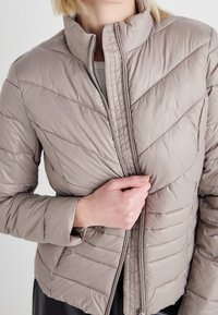 Lightweight, padded puffer jacket in beige with diagonal quilted design, high collar, and full zip; smooth, glossy texture.