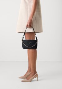 Czarna torebka z tłoczonym wzorem, srebrnym łańcuszkiem i trójkątnym logo, zestawiona z jasną sukienką w kratę i cielistymi szpilkami.