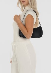 Zwarte geribbelde handtas met een gebogen vorm en een smalle schouderriem. Het materiaal heeft een zachte textuur, wat zorgt voor een modern, minimalistisch ontwerp.