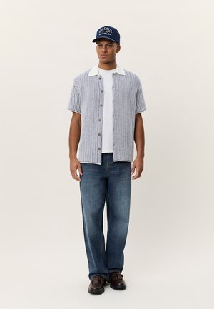 Πλεκτό πουκάμισο με κοντά μανίκια σε μπλε και λευκό, με λευκό γιακά και καφέ κουμπιά. Συνδυάζεται με ανοιχτόχρωμα μπλε τζιν και σκούρα παπούτσια. Φοριέται μπλε καπέλο.