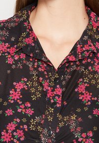 Chemise noire à col avec imprimé floral rose, jaune et rouge, boutonnée à l'avant, portée par une personne visible du cou à la partie supérieure de la poitrine.