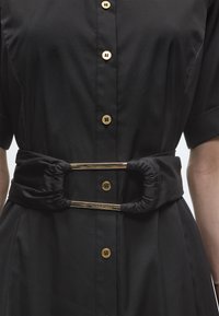 Μαύρο φόρεμα με κοντά μανίκια, χρυσά κουμπιά στο μπροστινό μέρος και φαρδιά μαύρη ζώνη με δύο χρυσές ορθογώνιες μεταλλικές διακοσμήσεις.