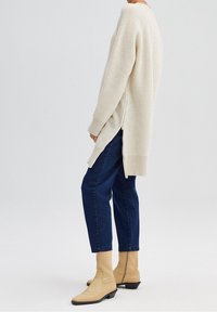 Cardigan lavorato a maglia color crema con spacchi laterali, abbinato a jeans blu scuro e stivaletti beige con punta squadrata e tacco basso.